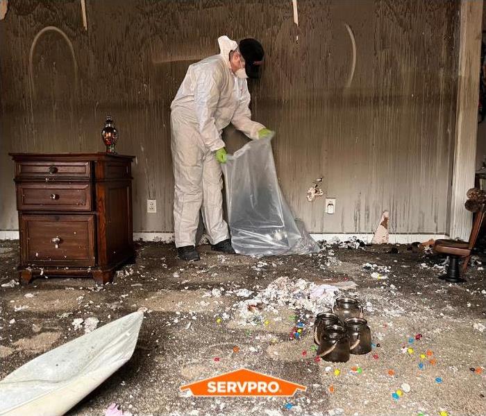 SERVPRO technician cleaning up contents in a fire, in white suit, holding white trash bag, a dark brown wall of soot behind